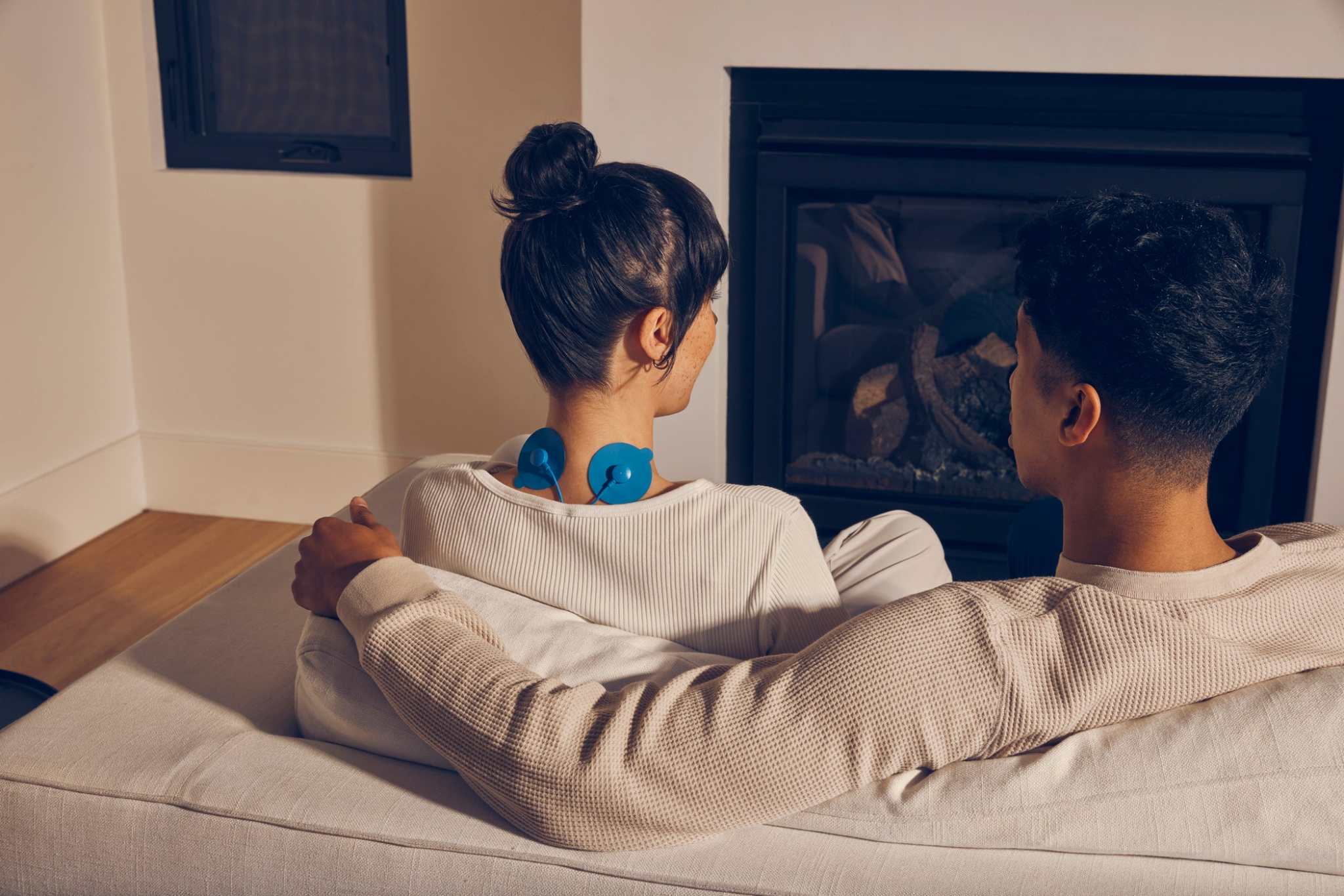 couple using PowerDot Uno app controlled muscle stimulator pads on neck while sitting on couch near fireplace