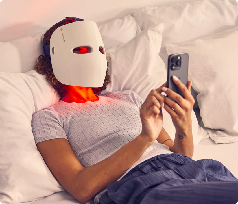 Woman lying on bed using TheraFace Mask cordless LED light therapy device for skin rejuvenation while holding smartphone