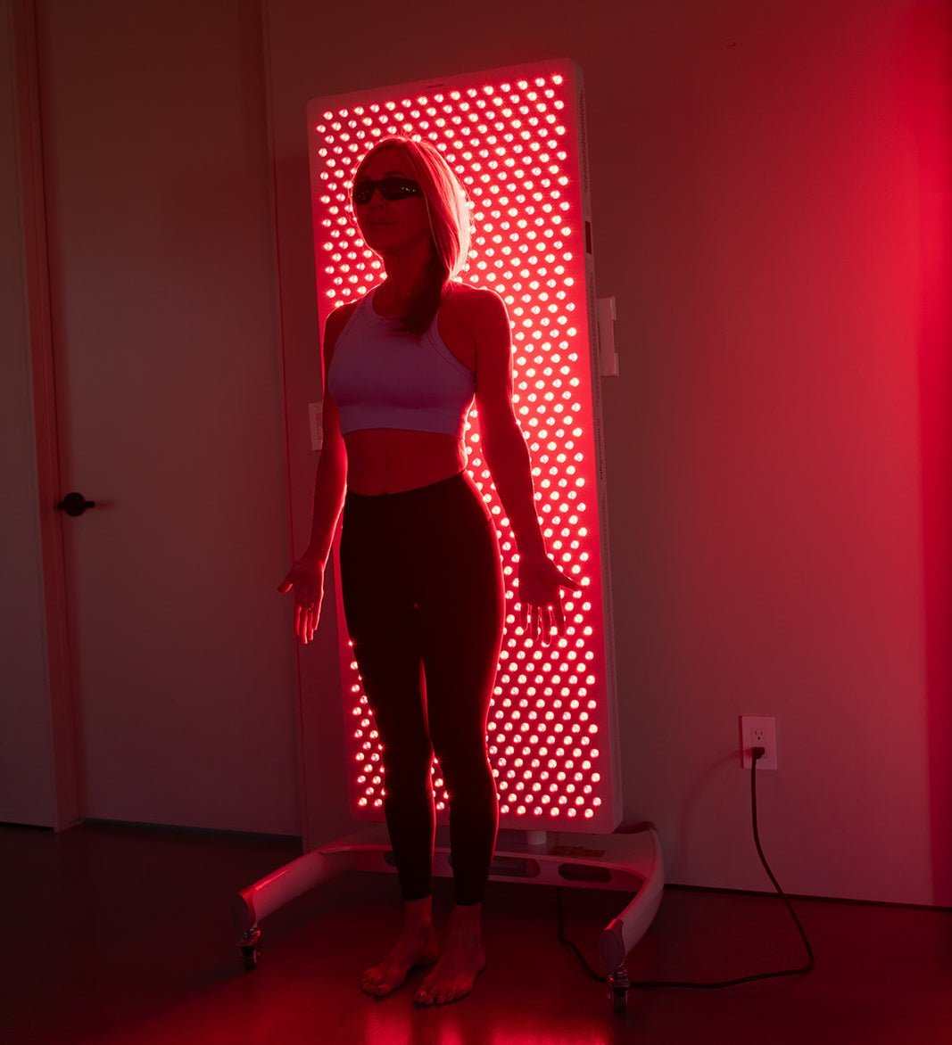 Woman using MitoPRO X red light therapy panel in dark room with protective glasses