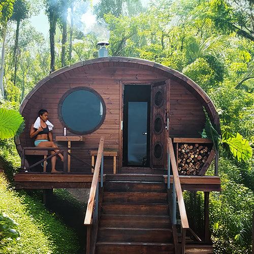 SaunaLife GL6 outdoor wooden barrel sauna kit in forest setting with woman sitting on porch