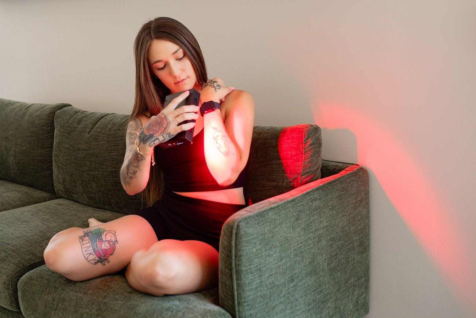 Woman using Mito Mobile portable red light therapy device on arm while sitting on couch