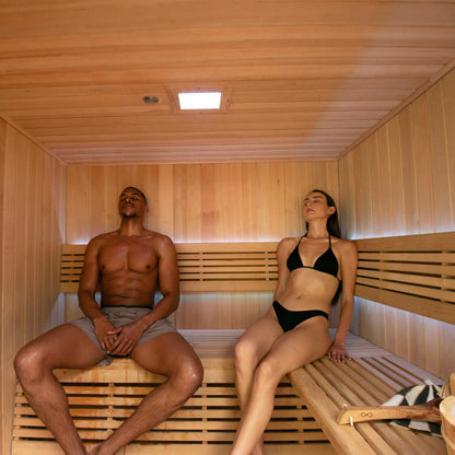 6 Person Traditional Sauna interior with two people relaxing on wooden L-bench seating, natural Hemlock wood finish, and ceiling light