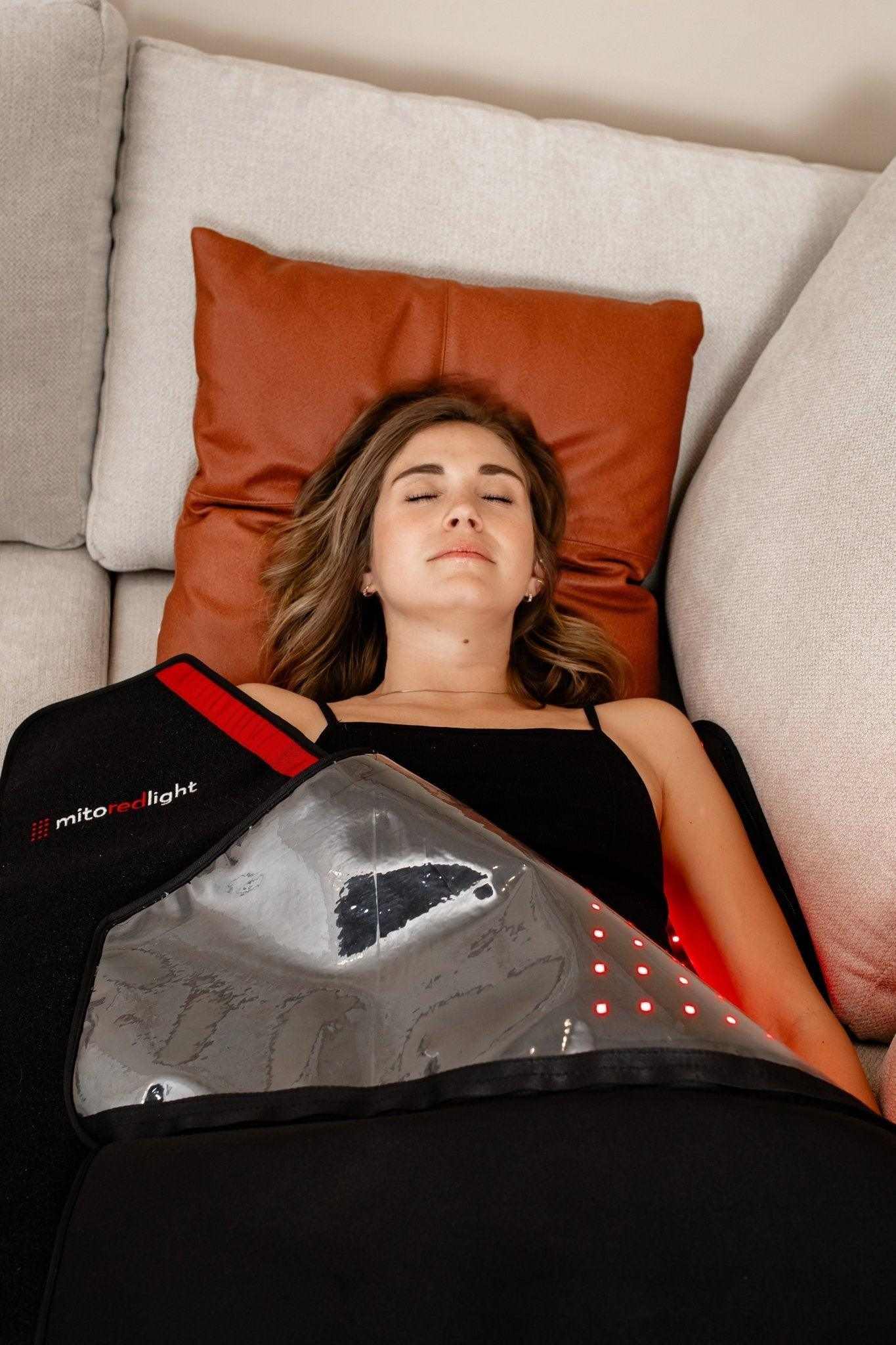 Woman using MitoPOD™ portable full body red light therapy device on couch